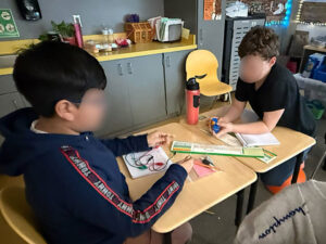 Two students sit facing each other with desks pushed together between them. The student on the right holds a blue D-cell battery holder and battery while leaning on a notebook. The student on the left holds the ends of two alligator test leads which are resting on a notebook on his desk.