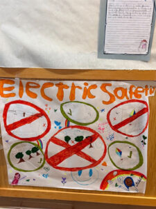 On a wall a page with a paragraph of young student writing is hung on a white paper. Below the white paper wall is a wooden wall with a hand drawn poster. The poster says "Electric Safetiy" in orange paint. Below the title are six circled images. In the top left with a single red cross bar through the red circle is a drawing of an item stuck in powerlines and clouds. In the upper middle is a green cicle with a green checkbox in the circle is a stick person standing away from the power lines. The right top circle is red with a red corss bar through it. This image shows a kite with power lines. In the bottom left is a green circule with three green trees. In the middle is a red circle with a full red X There are three trees, powerlines, and clouds in the circle. In the bootom right is a green circle with a yellow, red, blue, and dark blue kite flying among clouds. In the bottom left and right corners are two self portraits of girls with their names. Writing is not clear in the photo.