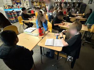 A view of a classroom. Students sit in small groups of 2 or 3 with their desks pushed together facing each other. Students have alligator test leads on desks in each group. Some students have open notebooks on their desk.