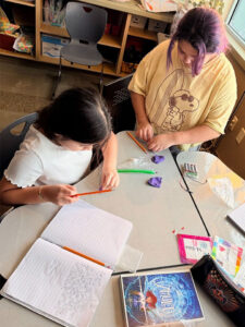 Two students work at a triangle shaped desk pushed into a group of four. On the desk is a book (Amulet) an open notebook with half a page of student writing (can not be read in image). The student sitting at the desk holds a red stick over some purple scultping clay. The student standing behind the desk at the top of the photo has two lumps of purple clay and is manipulating an item that can't be seen. Next to her hands is battery holder with 4 AA batteries and two wires.