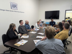Teachers sit around a conference table. On the wall is the logo for Constructing Hope underneath the logo is reads "Oregon State Senator Robert Boyer Boardroom" A TV is mounted on the far wall and currently off.