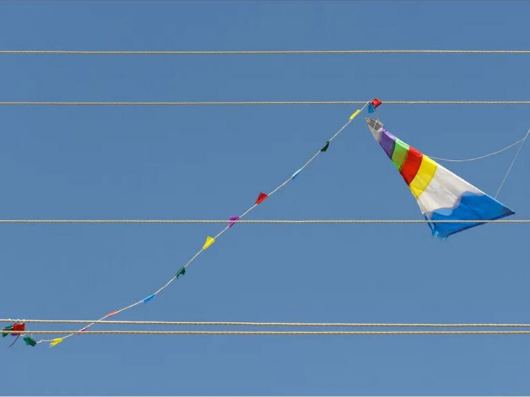 A blue, white, yellow, and yed stripped kite is tangled in a powerline against a blue sky. The tail of the kite has little flags in the same colors trailing down the wires to the left.