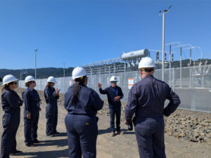 Teachers stand facing away from the camera wearing dark blue fire safe jumpsuits and white hardhats. In front of the gorup a man in a hardhat stands before a fenced off high voltage transfer station.