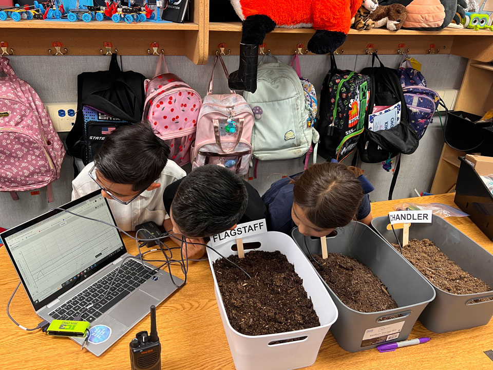 Three students stand behind a table and look away from the camera toward an open laptop screen on the table. The laptop shows a line graph with three data lines. In front of the studnts are three tubs of soil. One white and two grey. The leftmost (white) has a popsickle stick with the lable Flagsaff, the middle has a popsickle but the label can not be seen, the right most says Chandler. All tubs have a black wire coming out connected to the laptop.