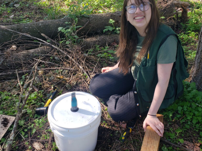 A girl sits on the dirt in a natural area holding a wooden plank on the ground, in front of her is a white bucket driven into the soil with lid and a Vernier Go Direct CO2 sensor in the top. A rubber mallet can be seen behind the bucket.